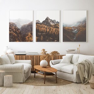 May include: Three framed wall art prints depicting mountain landscapes with golden autumn forests, rocky peaks, and misty valleys. The artwork is displayed above a wooden sideboard, two white upholstered sofas, and a wooden oval coffee table.