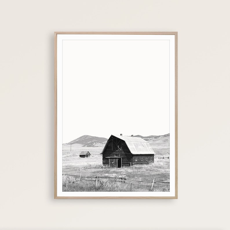 May include: A black and white photograph of a wooden barn with a metal roof in a field. The barn is surrounded by a wooden fence and there are rolling hills in the background.