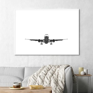 May include: Monochrome framed print of an aeroplane in flight, viewed head-on. The aircraft is centered against a bright white background, with its landing gear extended. The print is displayed above a sofa.