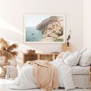 May include: A white bed with a tan throw blanket and white pillows. A framed photograph of a coastal town with colorful buildings on a cliffside overlooking the ocean is hanging on the wall above the bed.