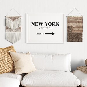 May include: A framed rectangular wall art print featuring the black text NEW YORK, NEW YORK, and 2908 MI with a right-pointing arrow. Flanking the print are two woven wall hangings, one with white fringe and one with brown textured patterns.