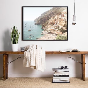 May include: A framed photograph of a coastal town with colorful buildings on a hillside overlooking a blue ocean. The photo is hanging on a white wall above a wooden bench with a white blanket, a stack of books, and a pair of sunglasses.