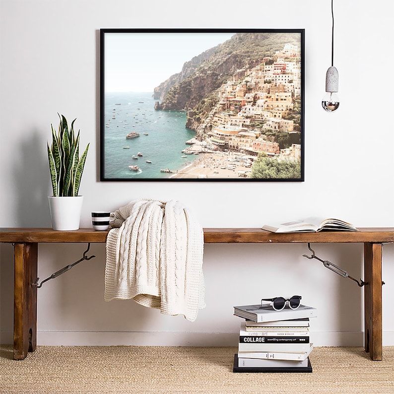 May include: A framed photograph of a coastal town with colorful buildings on a hillside overlooking a blue ocean. The photo is hanging on a white wall above a wooden bench with a white blanket, a stack of books, and a pair of sunglasses.