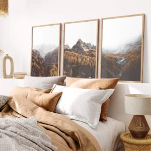 May include: Three vertical framed wall art prints depicting a mountain landscape with golden autumn trees, rocky peaks, and misty valleys. The prints are displayed above a bed with layered tan, white, and gray bedding and a textured table lamp.