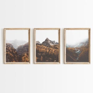 May include: A set of three vertical framed wall art prints featuring mountain landscapes. Each print displays a rocky mountain peak surrounded by dense forests with golden-brown autumn foliage, misty valleys, and a winding stream under a bright sky.