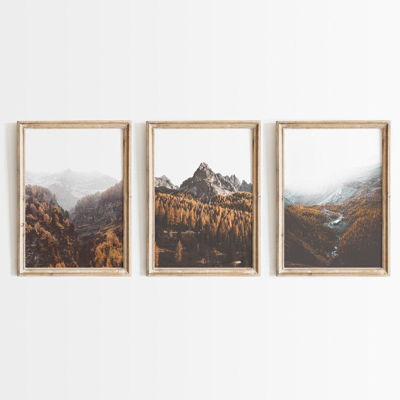 May include: A set of three vertical framed wall art prints featuring mountain landscapes. Each print displays a rocky mountain peak surrounded by dense forests with golden-brown autumn foliage, misty valleys, and a winding stream under a bright sky.