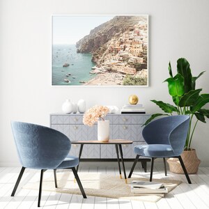 May include: Two blue velvet chairs with black legs and gold accents, facing each other across a wooden table with a vase of pink flowers. A framed photograph of a coastal town with a white frame hangs on the wall behind the chairs.
