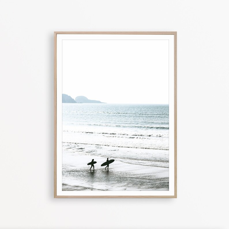 May include: Two people walk along a sandy beach carrying surfboards. The ocean is in the background with a distant mountain range.