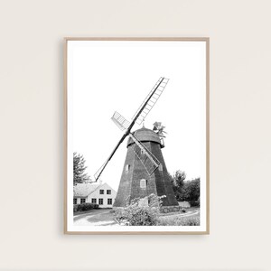May include: A black and white photograph of a windmill with a small house in the foreground. The windmill has wooden blades and a thatched roof. The house is a simple structure with a white facade and a few windows.