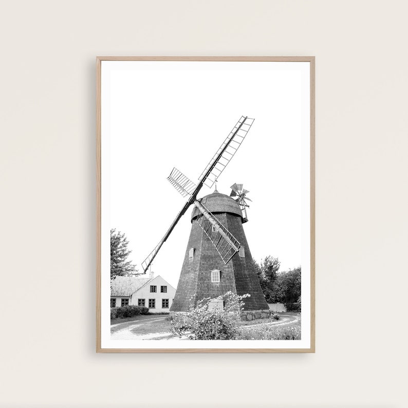 May include: A black and white photograph of a windmill with a small house in the foreground. The windmill has wooden blades and a thatched roof. The house is a simple structure with a white facade and a few windows.
