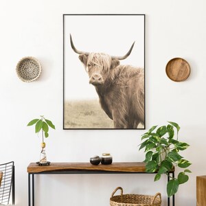 Może przedstawiać: Oprawiony wydruk przedstawiający krowę rasy Highland z długim, brązowym futrem i dużymi rogami. Grafika jest eksponowana na białej ścianie, uzupełniona drewnianym stołem konsolowym, elementami dekoracyjnymi i roślinami, tworząc rustykalną estetykę.