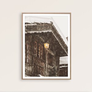 May include: A framed print of a snow-covered wooden building with a warm, glowing lantern. The building has multiple windows and a steeply pitched roof. Snow is falling, creating a winter scene. The frame is a light brown color.