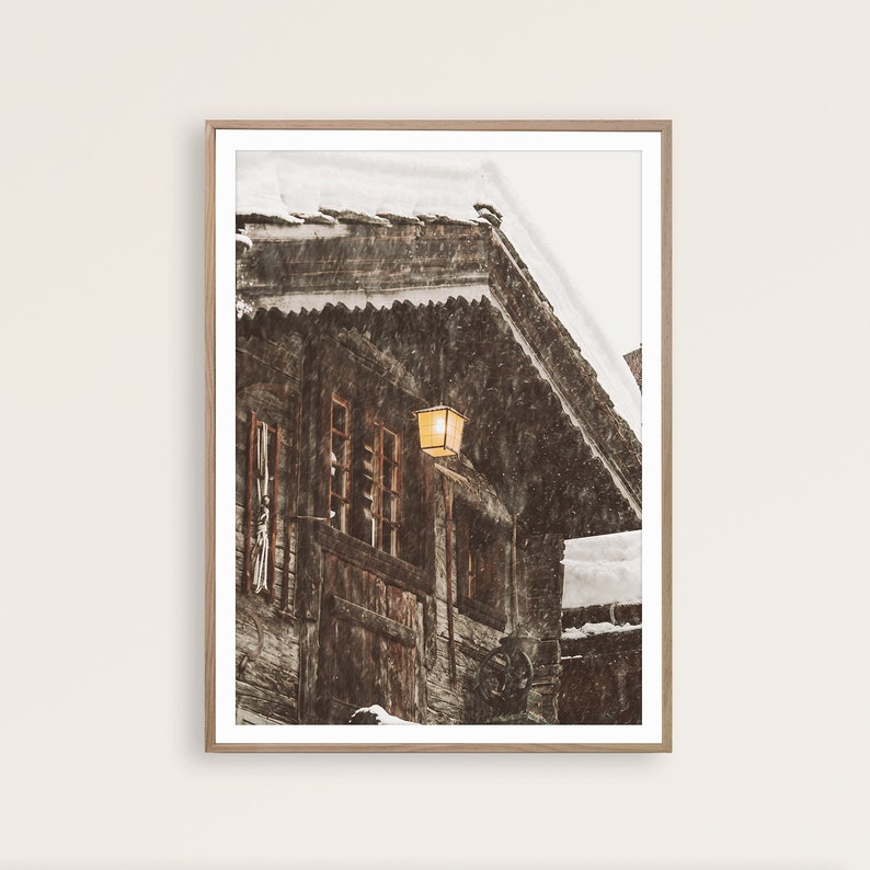 May include: A framed print of a snow-covered wooden building with a warm, glowing lantern. The building has multiple windows and a steeply pitched roof. Snow is falling, creating a winter scene. The frame is a light brown color.