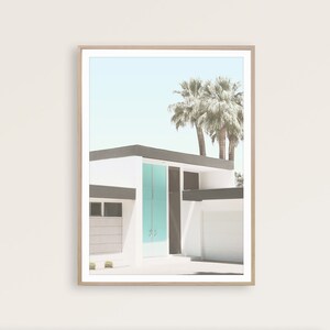 May include: A framed photograph of a modern, white house with a teal blue door. The house has a flat roof and a palm tree in the background. The photo is in a light wood frame.
