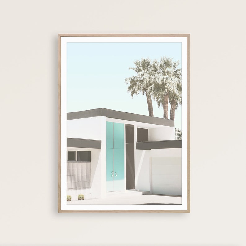 May include: A framed photograph of a modern, white house with a teal blue door. The house has a flat roof and a palm tree in the background. The photo is in a light wood frame.