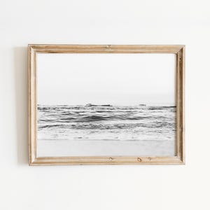 May include: A rectangular wooden frame containing a black and white photograph of ocean waves rolling toward a sandy shore. The frame has a light brown, natural wood grain finish and surrounds the horizontal landscape image of the sea.