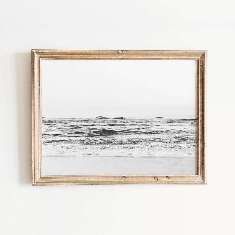 May include: A rectangular wooden frame containing a black and white photograph of ocean waves rolling toward a sandy shore. The frame has a light brown, natural wood grain finish and surrounds the horizontal landscape image of the sea.