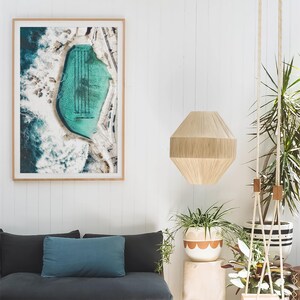 K&ouml;nnte beinhalten: Ein gerahmtes Luftbild eines t&uuml;rkisfarbenen Ozeanbeckens mit schwarzen Bahnenmarkierungen. Das Kunstwerk h&auml;ngt &uuml;ber einem dunkelgrauen Sofa mit einem tealfarbenen Kissen. Eine beige geometrische Pendelleuchte h&auml;ngt von der Decke, und Topfpflanzen f&uuml;gen Gr&uuml;n hinzu.