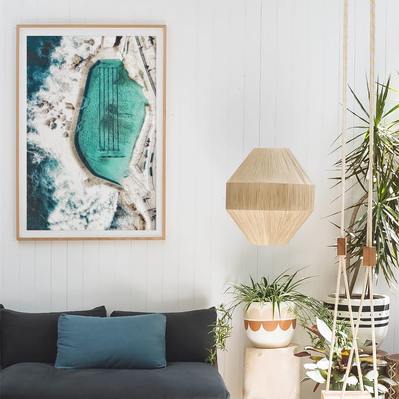 K&ouml;nnte beinhalten: Ein gerahmtes Luftbild eines t&uuml;rkisfarbenen Ozeanbeckens mit schwarzen Bahnenmarkierungen. Das Kunstwerk h&auml;ngt &uuml;ber einem dunkelgrauen Sofa mit einem tealfarbenen Kissen. Eine beige geometrische Pendelleuchte h&auml;ngt von der Decke, und Topfpflanzen f&uuml;gen Gr&uuml;n hinzu.