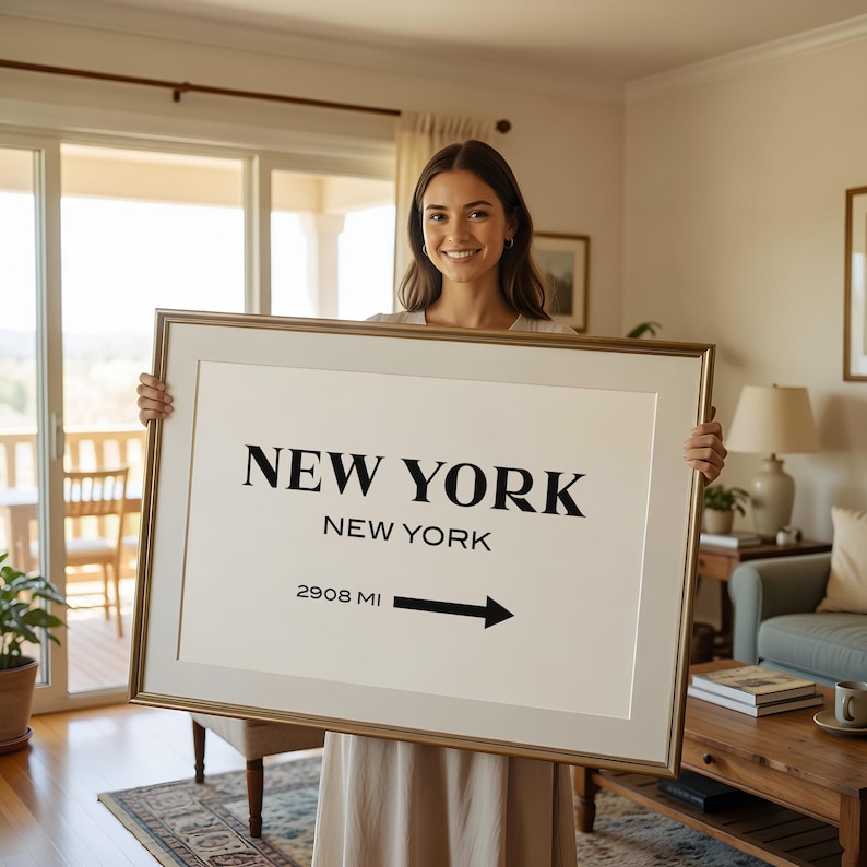 May include: A rectangular framed wall art piece featuring a white background with black text that reads NEW YORK, NEW YORK, and 2908 MI with a black arrow pointing to the right. The frame is a metallic gold color with a wide off-white matting border.