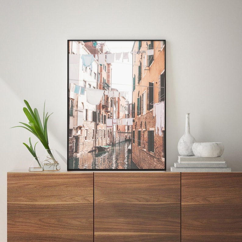 May include: A black and white photo of a narrow canal in Venice, Italy. Buildings line the canal, with laundry hanging from lines strung across the street. A small boat is in the canal.