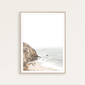Puede incluir: Una fotograf&iacute;a enmarcada de un acantilado rocoso que se encuentra con una playa de arena con olas espumosas rompiendo en la orilla. El oc&eacute;ano es de color azul claro y el cielo es de un blanco brumoso.