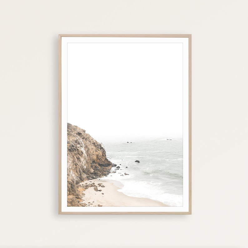 Puede incluir: Una fotograf&iacute;a enmarcada de un acantilado rocoso que se encuentra con una playa de arena con olas espumosas rompiendo en la orilla. El oc&eacute;ano es de color azul claro y el cielo es de un blanco brumoso.