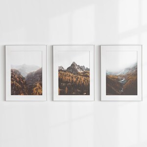 May include: Three vertical framed wall art prints featuring mountain landscapes. Each print displays a rocky peak surrounded by dense forests with golden autumn foliage, misty valleys, and a winding stream, all encased in simple white frames.