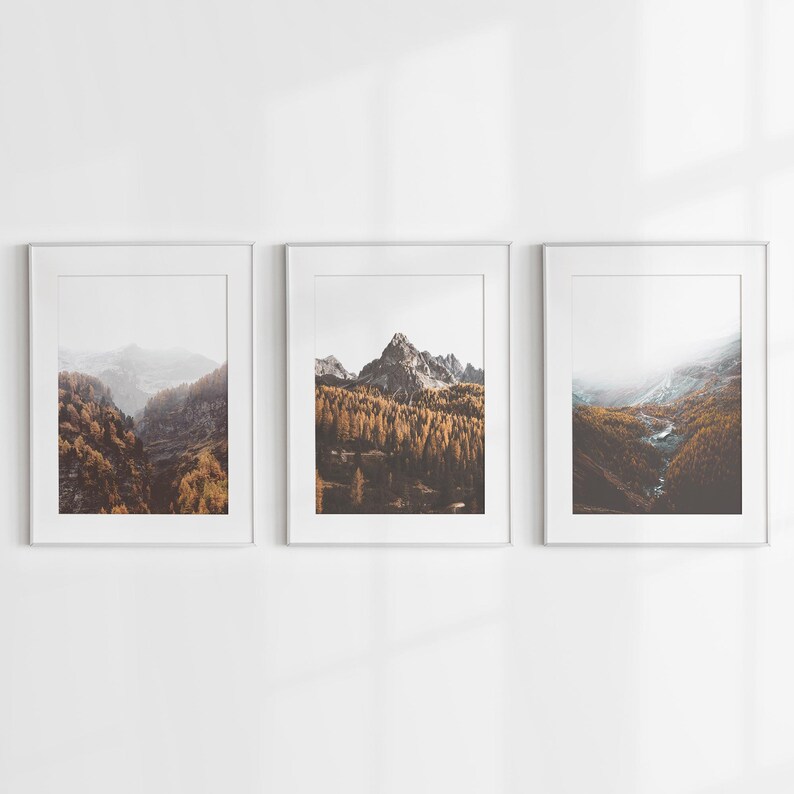 May include: Three vertical framed wall art prints featuring mountain landscapes. Each print displays a rocky peak surrounded by dense forests with golden autumn foliage, misty valleys, and a winding stream, all encased in simple white frames.