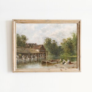 May include: A framed oil painting depicting a rural scene with a thatched-roof building, a river, and figures washing clothes. The painting is set against a cloudy sky and features lush green trees.