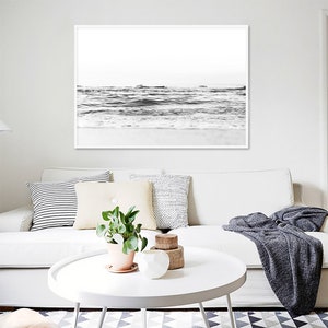 May include: A framed black and white landscape print of ocean waves crashing against a sandy shore, displayed in a thin white frame. The artwork features a high-contrast view of the sea with a bright, minimalist horizon line and soft foam.