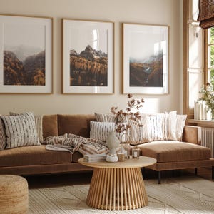 May include: A brown velvet sofa with striped throw pillows and a throw blanket, positioned in front of three framed mountain landscape prints. A round wooden coffee table with a slatted base sits in the center, holding a small vase and candles.