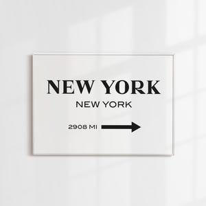 May include: A rectangular wall art print in a thin frame featuring the text NEW YORK in large black serif font, followed by NEW YORK in smaller sans-serif font, and the text 2908 MI with a black arrow pointing to the right on a white background.