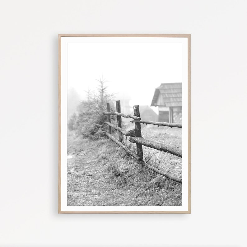 May include: A black and white photograph of a wooden fence in a foggy field. The fence is made of rough-hewn logs and is partially obscured by tall grass. A small wooden building is visible in the background.