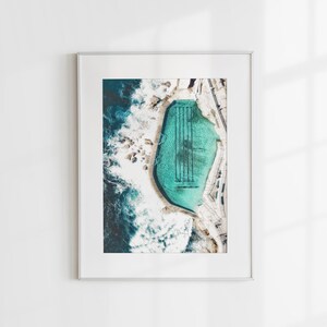 K&ouml;nnte beinhalten: Luftaufnahme eines t&uuml;rkisfarbenen Schwimmbeckens mit schwarzen Bahnen, eingebettet in eine felsige K&uuml;stenlinie mit tosenden Wellen. Das Bild ist in einem wei&szlig;en Rahmen eingefasst.