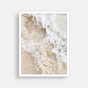 May include: A close-up of white foamy ocean waves crashing on a sandy beach. The sand is a light beige color and the waves are white and foamy.