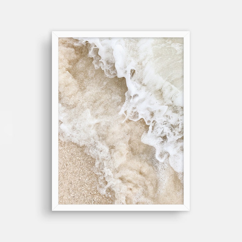 May include: A close-up of white foamy ocean waves crashing on a sandy beach. The sand is a light beige color and the waves are white and foamy.