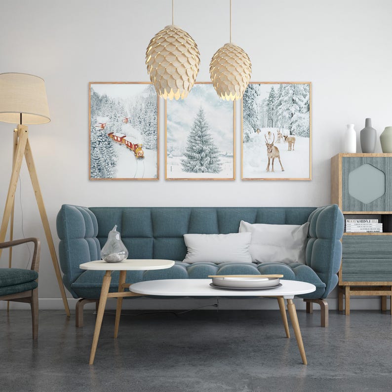 May include: A living room featuring a blue tufted sofa, two white oval coffee tables, and a floor lamp. Three framed winter landscape prints hang on the wall, depicting a train in snow, a pine tree, and reindeer. Two pinecone-shaped pendant lights hang above.