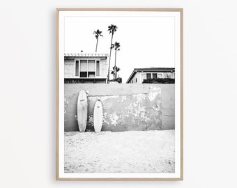 May include: Black and white framed print featuring two surfboards leaning against a weathered concrete wall on a sandy beach. Palm trees and buildings are visible in the background, creating a coastal scene.