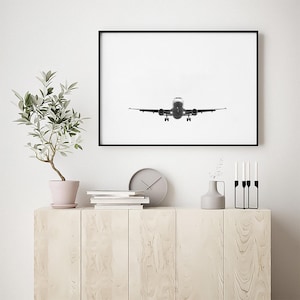 May include: A framed monochrome photograph of an aircraft in flight. The plane is centrally positioned, with its landing gear down, against a bright white backdrop. The photograph is displayed above a light-coloured wooden cabinet.