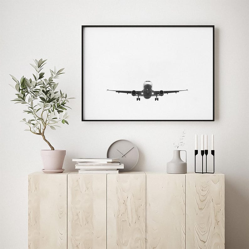 May include: A framed monochrome photograph of an aircraft in flight. The plane is centrally positioned, with its landing gear down, against a bright white backdrop. The photograph is displayed above a light-coloured wooden cabinet.