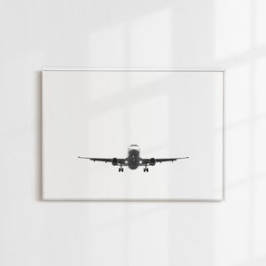May include: Monochrome framed print featuring an airplane in flight, seen from the front. The aircraft is centrally positioned, with its wings spread. The image is set against a white backdrop, offering a minimalist style. The plane's lights are on.