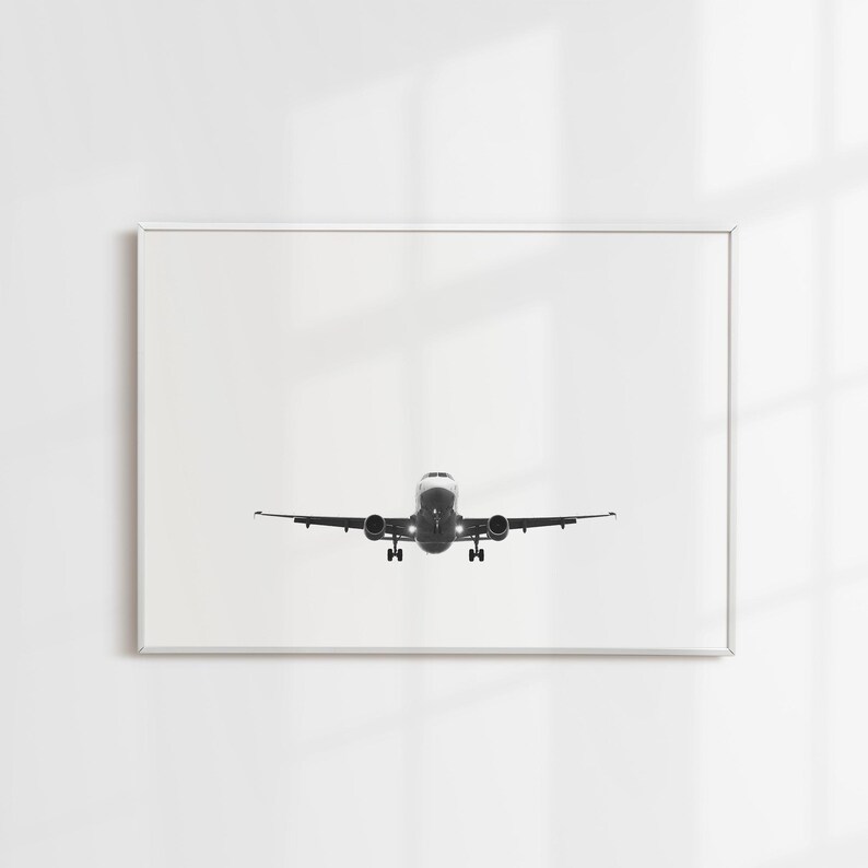 May include: Monochrome framed print featuring an airplane in flight, seen from the front. The aircraft is centrally positioned, with its wings spread. The image is set against a white backdrop, offering a minimalist style. The plane's lights are on.