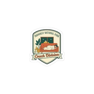 May include: A sticker with a shield design featuring the text "Indoorsy National Park" and "Couch Division." The image shows a person relaxing on a red couch, with a window, plant, and side table. The sticker has a white border.