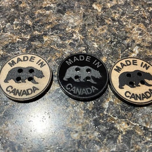 May include: Three round buttons with a black bear silhouette and the text "MADE IN CANADA". One button is black, one is brown, and one is tan.