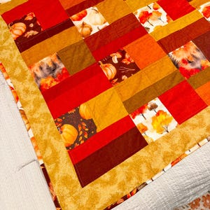 May include: A patchwork quilt featuring a repeating pattern of squares in shades of red, orange, yellow, and brown. The fabric squares display prints of pumpkins, autumn leaves, and trees, bordered by a textured golden-yellow fabric trim.