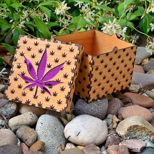 May include: A wooden box with a laser-cut cannabis leaf design and a purple leaf inlay. The box is open, revealing its interior. The box is surrounded by rocks and greenery.
