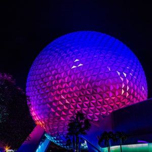May include: A large, geodesic dome-shaped building illuminated in a gradient of pink and blue at night. The building is made up of many small, triangular panels.