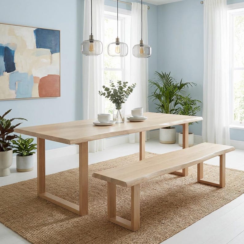 Pode incluir: Um conjunto de mesa de jantar e banco de madeira clara. A mesa tem um tampo retangular e uma base em forma de U. O banco tem um design semelhante. O conjunto est&aacute; sobre um tapete de juta tecido numa sala com paredes azul claro. A mesa tem cerca de 180 cm de comprimento.