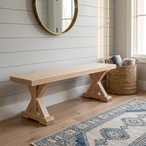 May include: A light-coloured wooden bench with an X-shaped base. The bench is approximately 152 cm long. A round gold mirror hangs on the wall above the bench. A woven basket with rolled towels sits nearby.